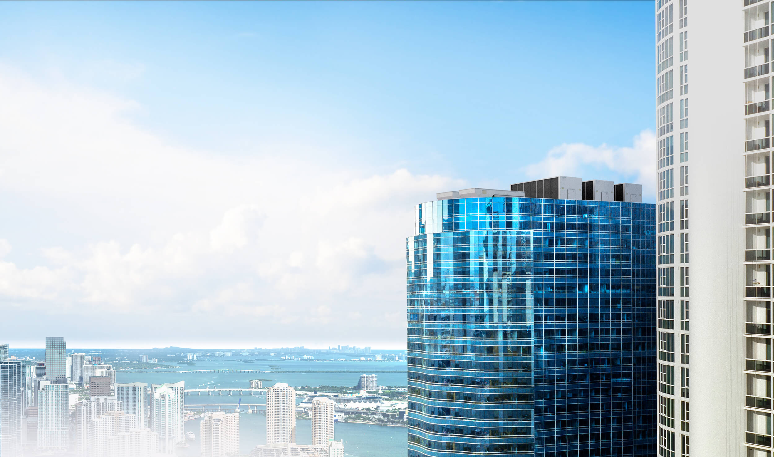 Tall blue-glass skyscraper on the right with a neighboring white high-rise against a clear blue sky, part of a city skyline.